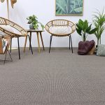 A cozy, minimalist living space featuring two rattan chairs with cushions, a small table, potted plants, and a textured carpet.