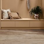 A cozy nook with a beige bench, pillows, plant, and wood-look laminate flooring.