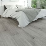 A cozy bedroom with light gray laminate flooring, a neatly made bed, and natural light from large windows.