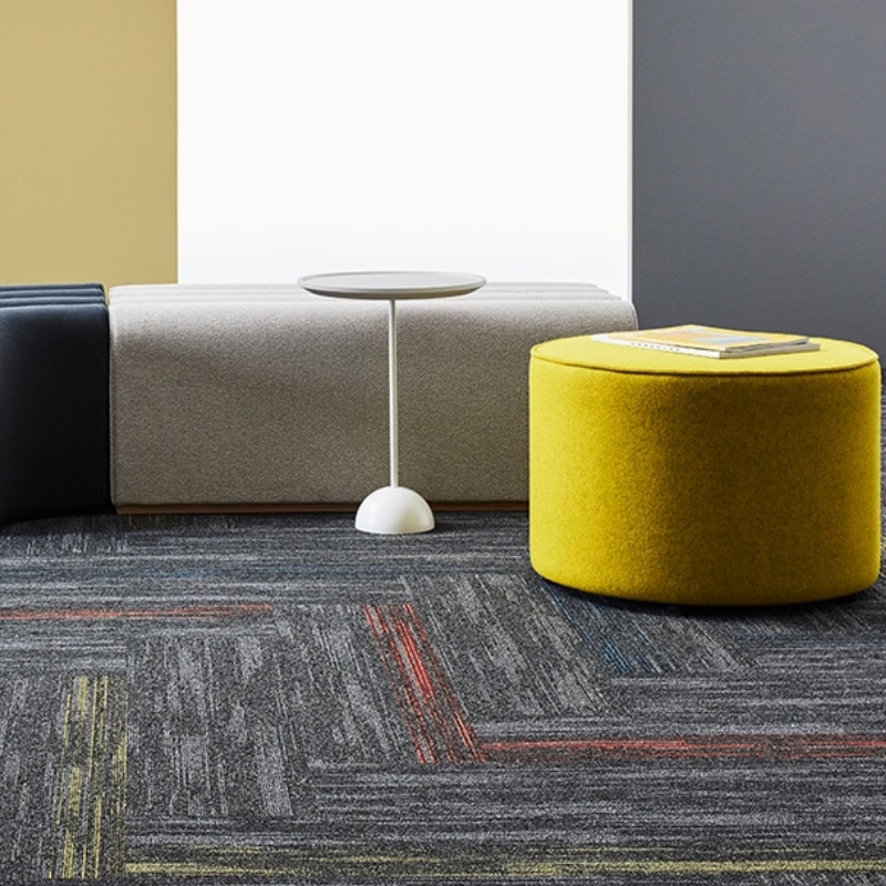A modern lounge area with colorful carpet tiles, a yellow ottoman, and minimalist furniture.