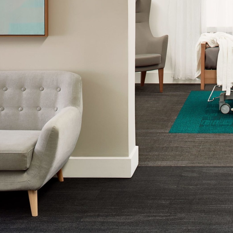 A modern living room with a comfortable sofa, a chair, and a bed in the background. The floor is covered with a dark, patterned carpet.
