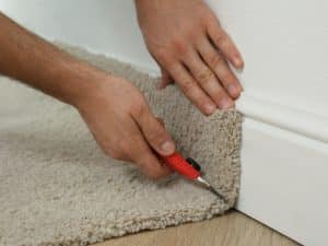 A close-up of hands trimming beige carpet with a utility knife during a DIY carpet installation job, against a white wall and skirting board.