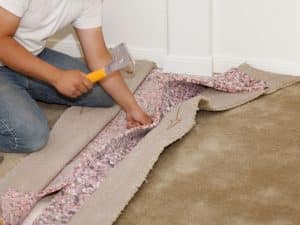 Man repairing carpet seams using hammer and padding during carpet installation on beige floor near white baseboard.